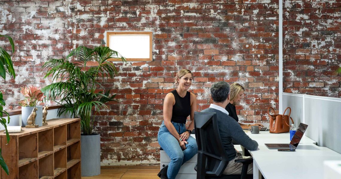 Three coworkers casually chatting near desks at ko&co workhub, surrounded by indoor plants and exposed brick walls. South Melbourne and Port Melbourne
