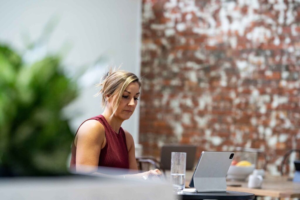 A woman works on her tablet at a bar-height table at ko&co workhub, with a glass of water nearby and an exposed brick wall in the background. South melbourne coworking