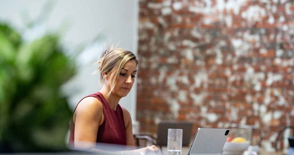 A woman works on her tablet at a bar-height table at ko&co workhub, with a glass of water nearby and an exposed brick wall in the background. South melbourne coworking